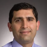 Shahin Nudehi, Ph.D., P.E., a man with short dark hair wearing a light blue collared shirt, smiles slightly against a dark background.