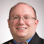 A smiling man with glasses, Nicholas S. Rosasco is dressed in a suit, blue shirt, and patterned bow tie, standing against a plain background.