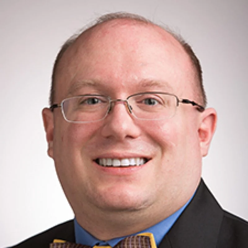 A smiling man with glasses, Nicholas S. Rosasco is dressed in a suit, blue shirt, and patterned bow tie, standing against a plain background.