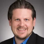 Smiling man with short brown hair and beard, wearing a blue shirt and black blazer, stands against a gray background—meet George Potter.