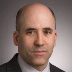 Paul Lapsansky, a man in a suit with a shaved head, poses for a professional headshot against a gray background.
