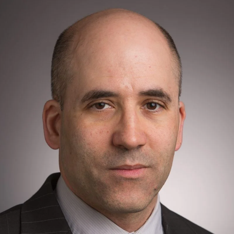 Paul Lapsansky, a man in a suit with a shaved head, poses for a professional headshot against a gray background.