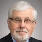 Older man with white hair, beard, glasses, and suit, smiling slightly against a neutral background.