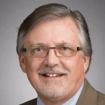 Smiling middle-aged man with glasses, gray hair, and a trimmed beard, wearing a tan suit jacket.