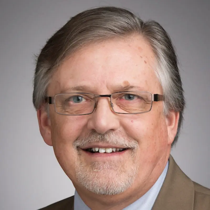 Smiling middle-aged man with glasses, gray hair, and a trimmed beard, wearing a tan suit jacket.
