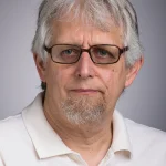 Older man with gray hair, glasses, a short beard, and a white polo shirt facing the camera—could be Michael Glass.