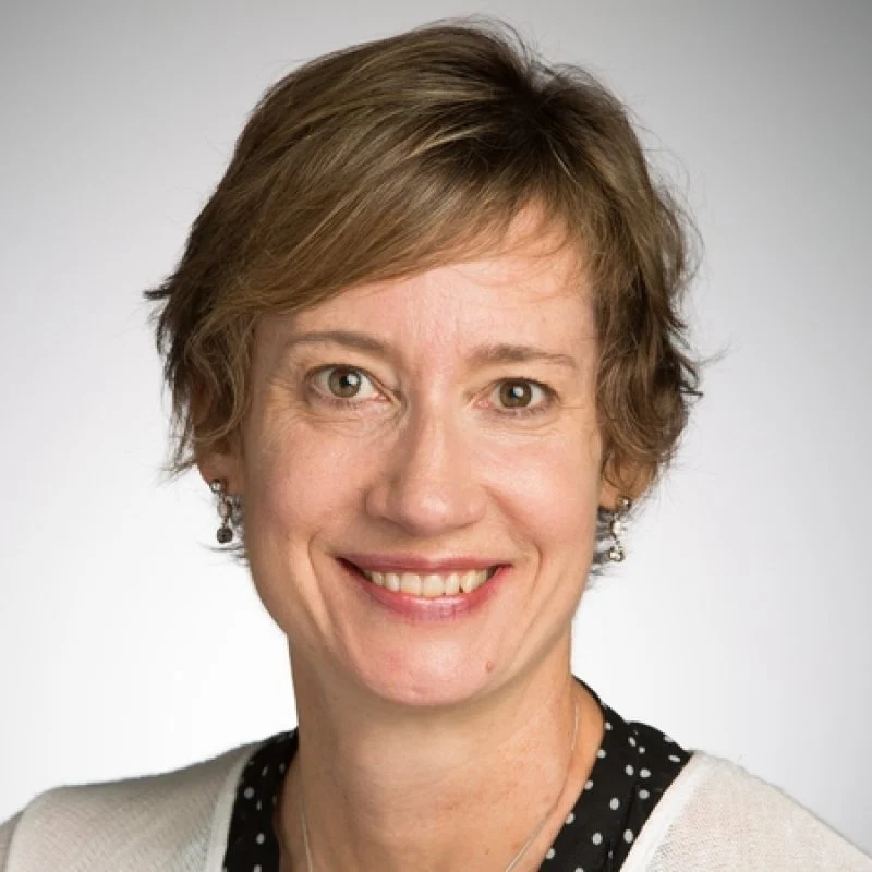 Smiling woman with short light brown hair, wearing a white top and earrings, poses against a plain background—Kelly Belanger.