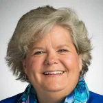 Smiling woman, Nola Schmidt, with short gray hair, wears a blue patterned scarf and blue jacket against a light background.