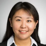 Woman with long dark hair smiling, wearing a white collared shirt and black blazer, on a light background.