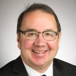 Smiling man wearing glasses, a suit, and a tie, with short dark hair against a light background—Jeffrey C. Brown.