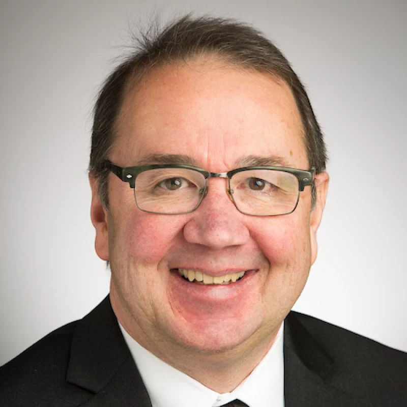 Smiling man wearing glasses, a suit, and a tie, with short dark hair against a light background—Jeffrey C. Brown.