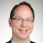 Smiling man with glasses and short brown hair, wearing a suit and checkered shirt, stands against a plain background—Gregg Johnson exudes confidence and professionalism.