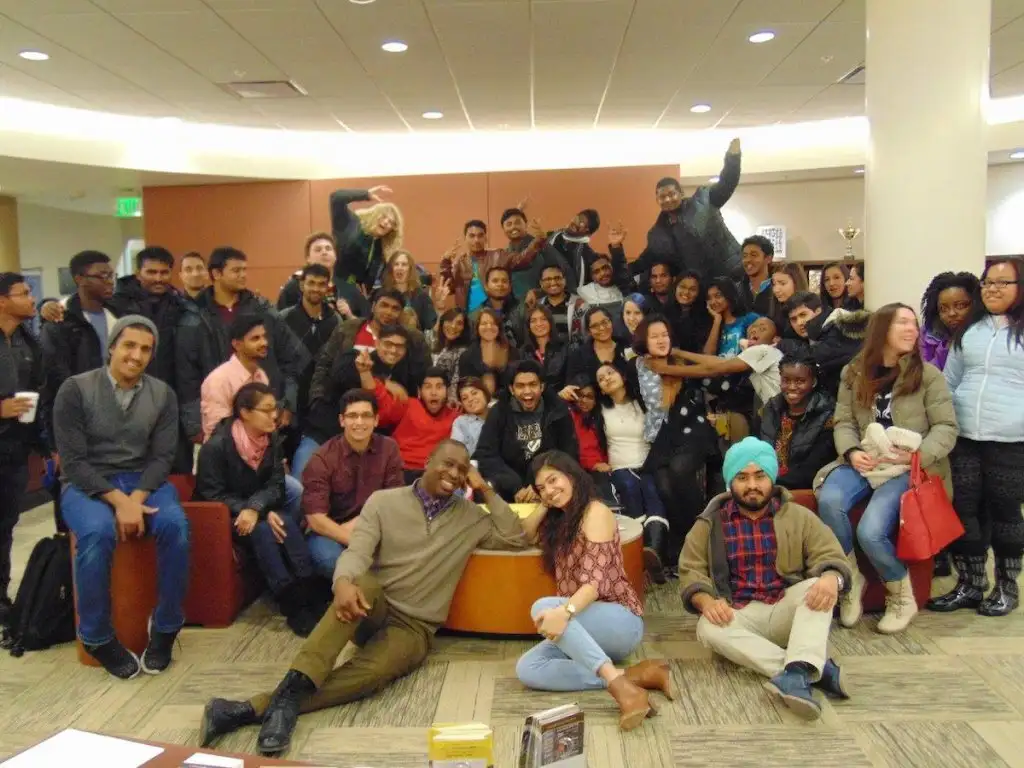 A large, diverse group of people pose together indoors, smiling and making playful gestures, showcasing the vibrant community supported by International Student Resources.
