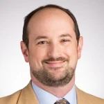 Smiling man with short brown hair and beard, wearing a tan jacket, blue shirt, and striped tie.