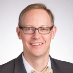 Smiling man with glasses, identified as Joseph Zaweski, wearing a dark suit, white shirt, and light-colored bow tie on a light background.