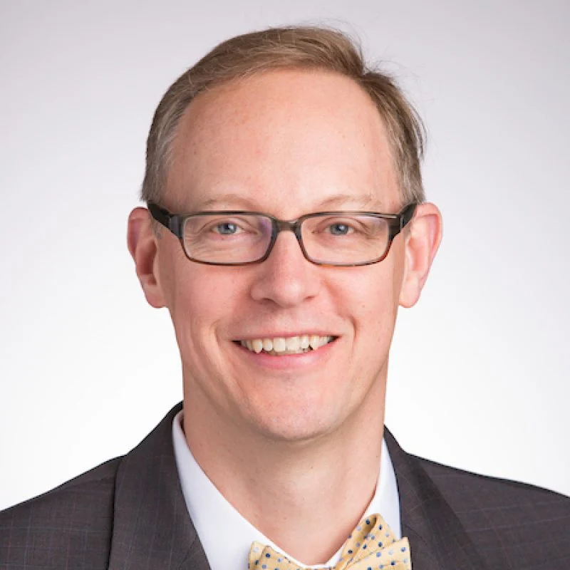Smiling man with glasses, identified as Joseph Zaweski, wearing a dark suit, white shirt, and light-colored bow tie on a light background.