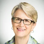 Smiling older woman with short blonde hair, glasses, and hoop earrings, wearing a patterned scarf—Randa Duvick.