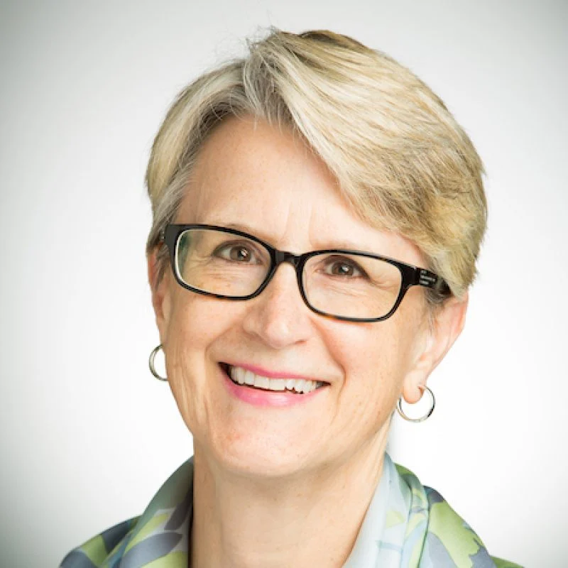 Smiling older woman with short blonde hair, glasses, and hoop earrings, wearing a patterned scarf—Randa Duvick.