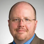Timothy Tomasik, a man with glasses, a short beard, and a suit, smiles slightly against a plain background.