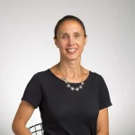 Agnes Howard, a woman with brown hair in a ponytail, wearing a black shirt and flower necklace, smiles warmly at the camera.
