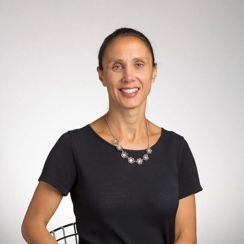 Agnes Howard, a woman with brown hair in a ponytail, wearing a black shirt and flower necklace, smiles warmly at the camera.