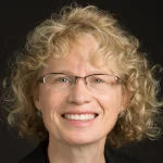 Smiling Elizabeth Burow-Flak, with curly blonde hair, glasses, and a black top, stands against a dark background.