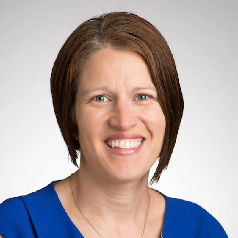 Smiling woman with straight brown hair in a blue top, facing the camera against a light background. Jennifer Hora.