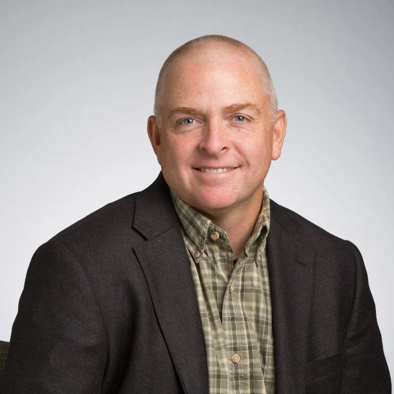 Smiling Thomas Albert Howard, a man with short hair in a plaid shirt and dark blazer, sits against a plain light background.