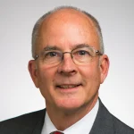 Smiling older man with glasses, short gray hair, and a gray suit against a plain light background. John Steele, MBA, exudes professionalism and confidence.