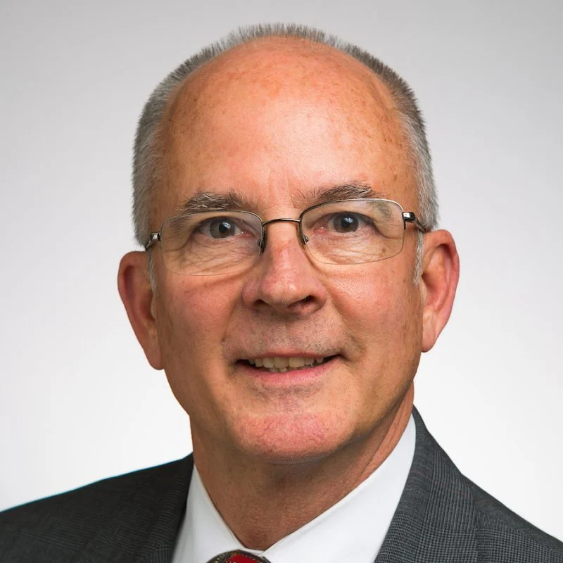 Smiling older man with glasses, short gray hair, and a gray suit against a plain light background. John Steele, MBA, exudes professionalism and confidence.