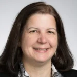 Anne Raich, Ph.D., a woman with long dark hair, smiles while wearing a patterned shirt and black jacket against a light background.