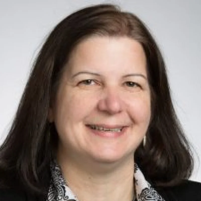 Anne Raich, Ph.D., a woman with long dark hair, smiles while wearing a patterned shirt and black jacket against a light background.