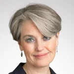 Smiling woman with short gray hair and light eyes, wearing earrings and a dark top against a light background—Maura Janton Cock.