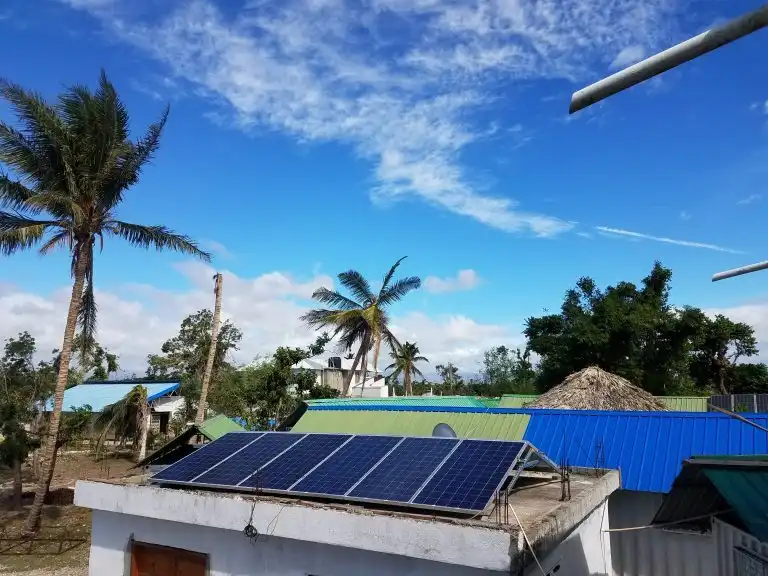 Solar panels on a rooftop capture energy beneath palm trees and blue sky, while gentle WAVES shimmer in the distance.