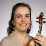 Katharina Uhde smiles while holding a violin, wearing pearl earrings and a necklace, against a light gray background.