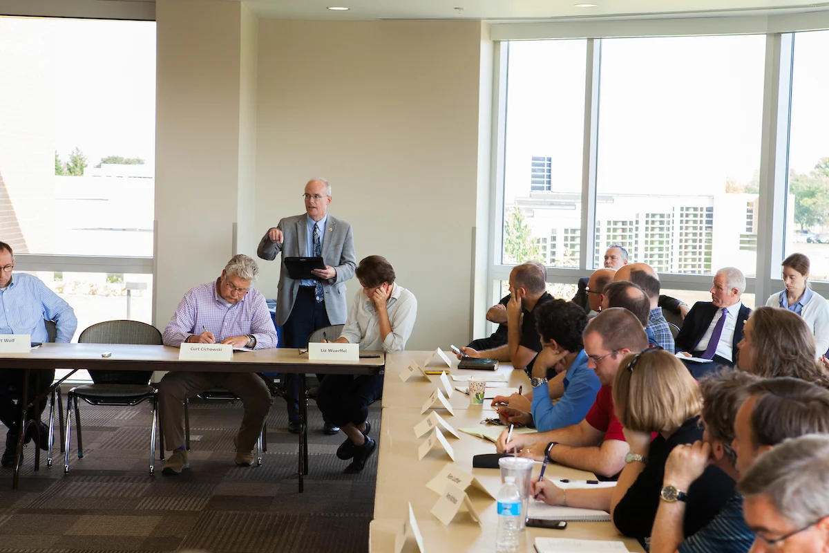 People sit at tables in a conference room while a man stands and speaks to the group about important academic policies.