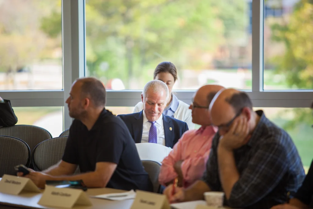 People seated at a table in a meeting room, some discussing items from the Faculty Calendar, with large windows in the background.