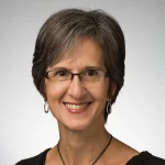 Smiling woman with short gray hair, glasses, hoop earrings, and a black top against a light background—Dawn Jeglum Bartusch.
