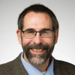 Smiling man with glasses, short dark hair, and a beard, wearing a brown jacket and blue shirt. Jay Grossman, P.E., Ph.D., exudes professionalism and warmth.