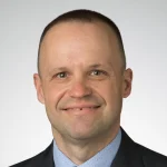Nicholas Denysenko, Ph.D., M.Div., smiles at the camera in a suit and tie against a plain light background.