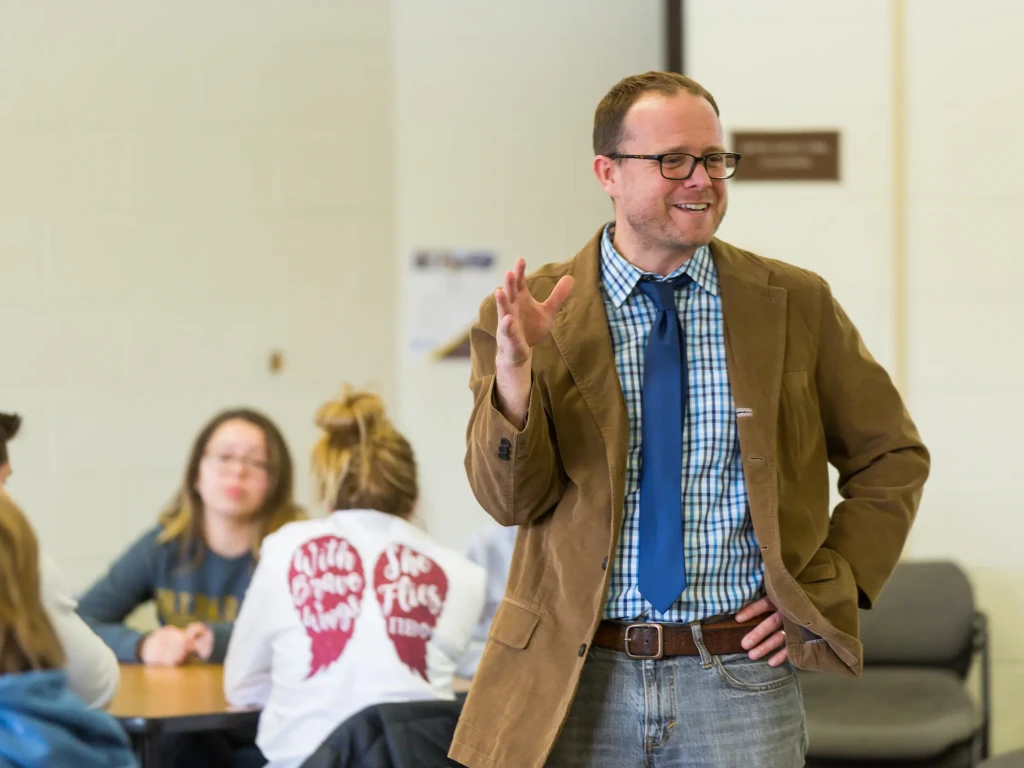 A man in a brown jacket and blue tie speaks to a group of academics seated at tables in a classroom.