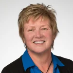 Smiling Lynette Rayman with short, light brown hair wears a blue shirt and black sweater against a white background.