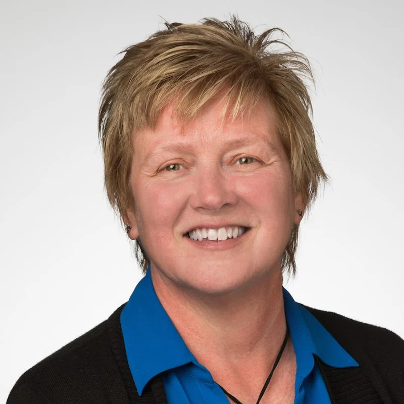 Smiling Lynette Rayman with short, light brown hair wears a blue shirt and black sweater against a white background.