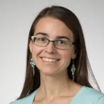 Bethany Luke, Ph.D., with long brown hair, glasses, turquoise earrings, and a light blue top, smiles at the camera.
