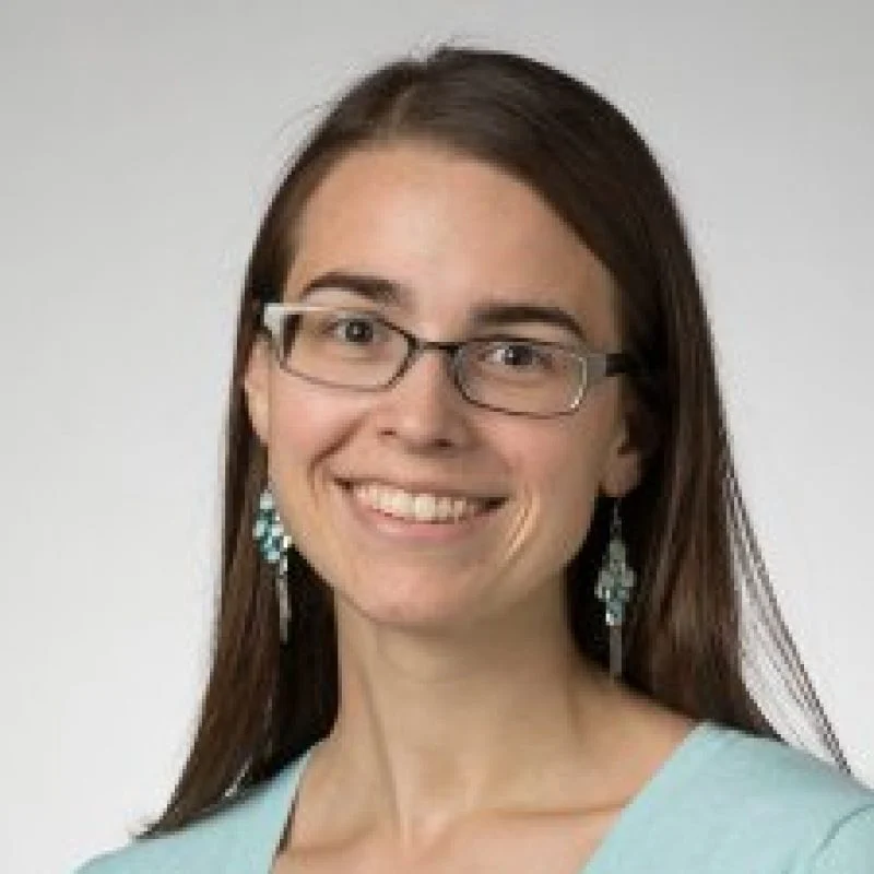 Bethany Luke, Ph.D., with long brown hair, glasses, turquoise earrings, and a light blue top, smiles at the camera.