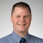Smiling man with short brown hair, Peter Krenzke, Ph.D., wearing a blue shirt and tie against a plain light background.