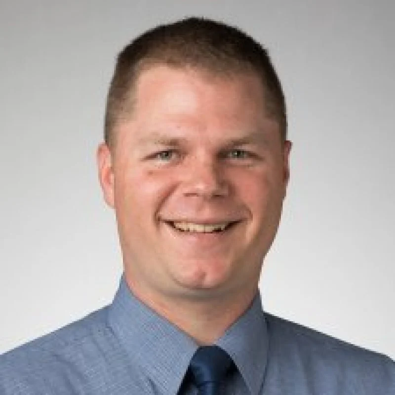 Smiling man with short brown hair, Peter Krenzke, Ph.D., wearing a blue shirt and tie against a plain light background.