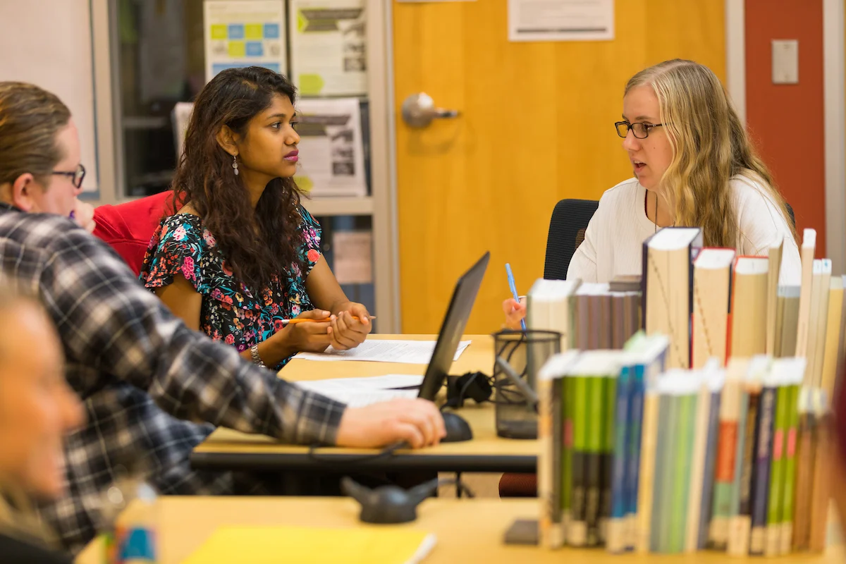 Three people sit at a library table talking, with books and a laptop in front of them, exploring student resources for their studies.
