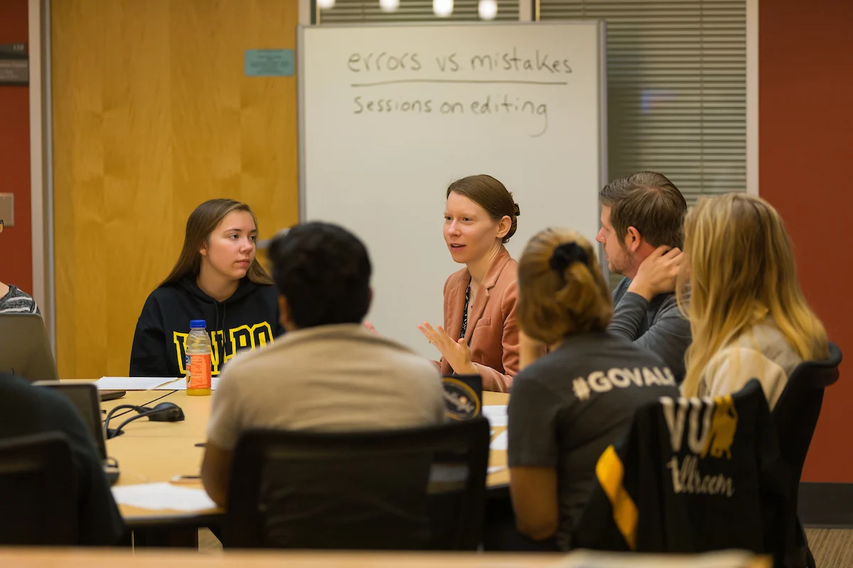 A group of people sit around a table, discussing in front of a whiteboard labeled “errors vs mistakes,” as they explore valuable Student Resources for improving their understanding.