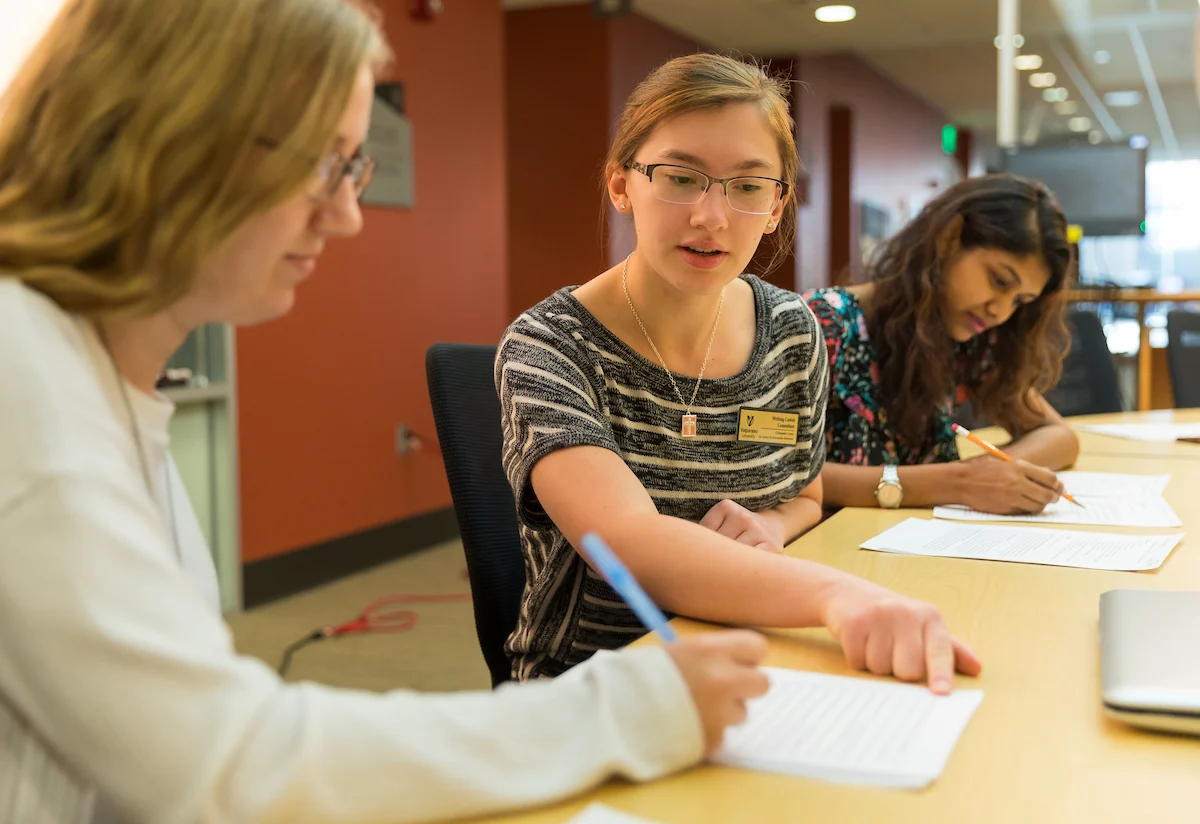 Three young women sit at a table, using student resources while writing on papers and discussing their work together.